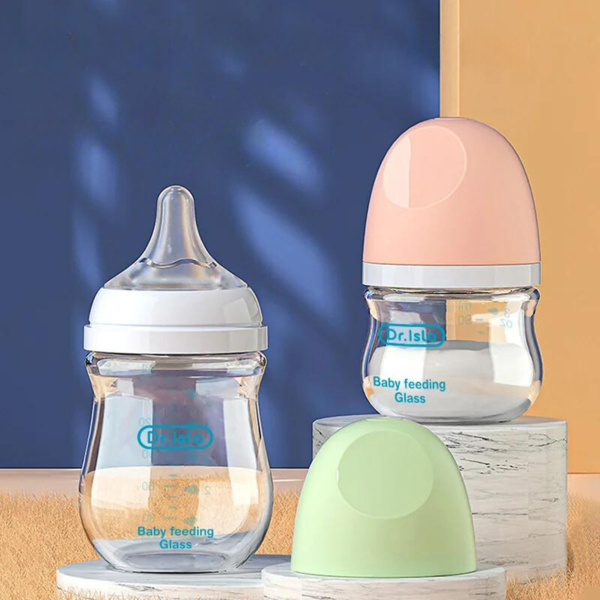 Three baby feeding bottles on display; one with a silicone nipple and pink cap, another with a light green cap placed on the side. The two bottles are labeled 'Dr. Isla Baby feeding Glass' against a colorful background.
