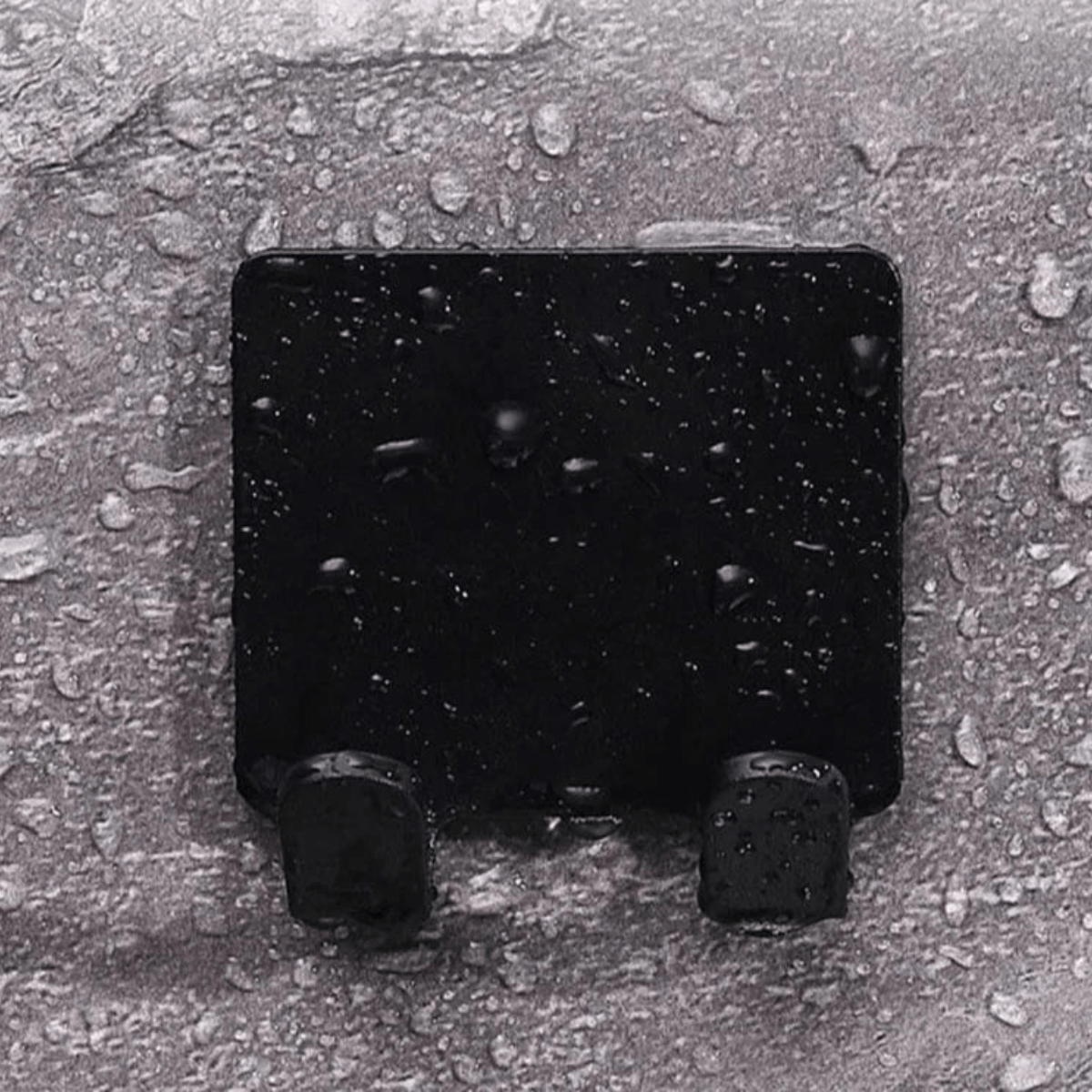 A black wall-mounted holder with two hooks, adhered to a wet, textured surface with water droplets scattered around.