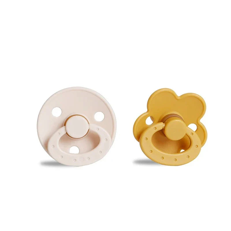 A round, beige pacifier with a ring handle next to a yellow, flower-shaped pacifier with a ring handle.