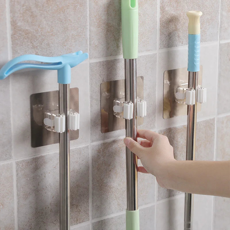 Three adhesive hooks are mounted on a tiled wall, each holding a mop. A person is adjusting one of the mops.